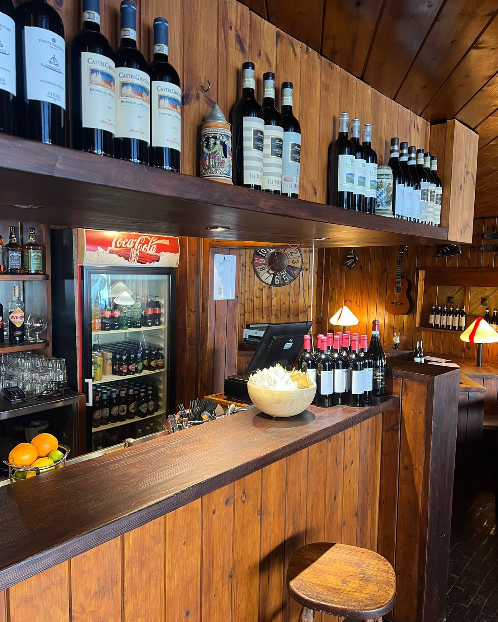 Bancone in legno con bottiglie di vino e snack, una ghiacciaia e qualche sgabello in un ambiente scarsamente illuminato.
