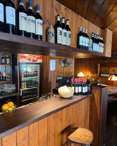 Bancone in legno con bottiglie di vino e snack, una ghiacciaia e qualche sgabello in un ambiente scarsamente illuminato.