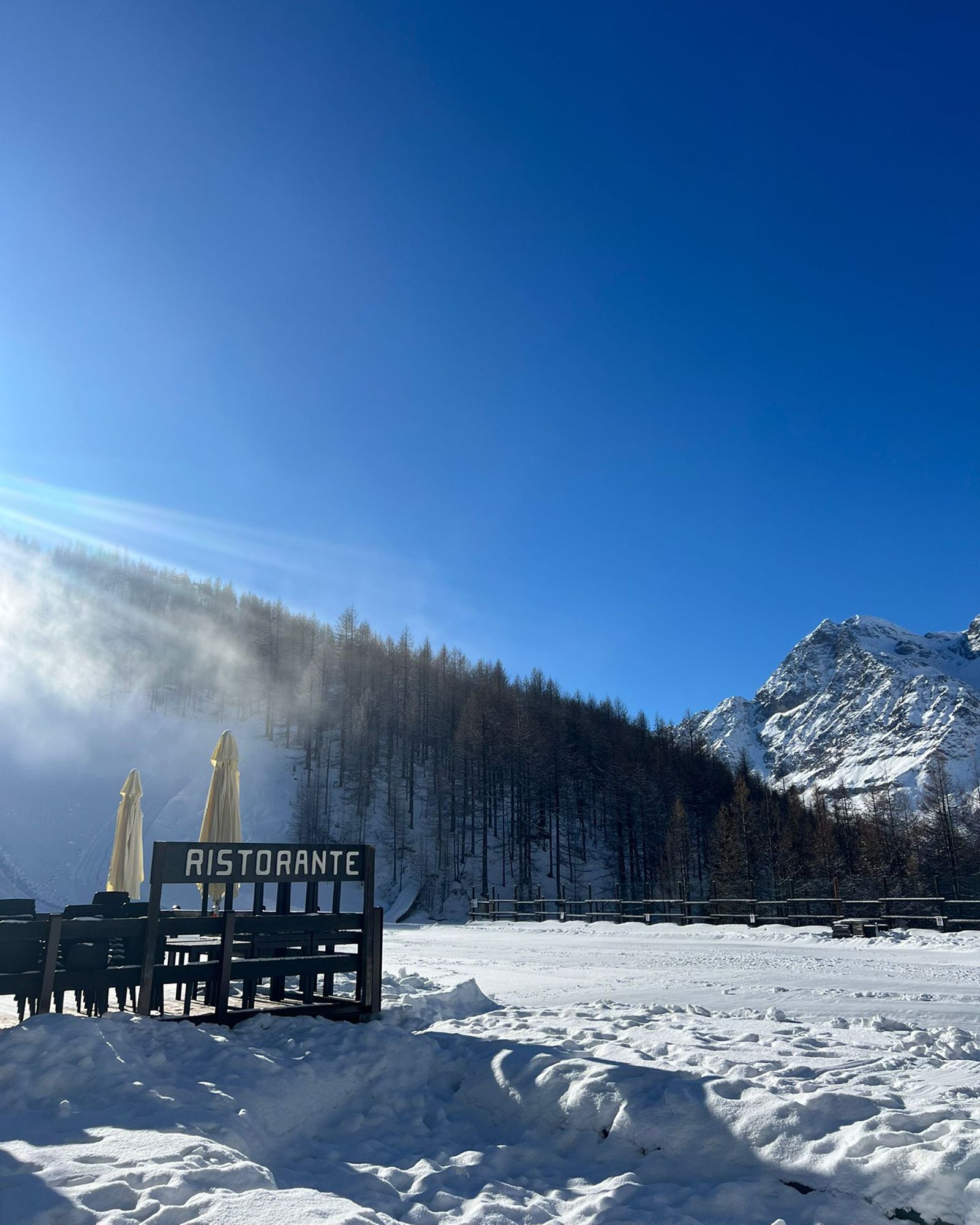 Paesaggio montano innevato con l'insegna di un ristorante, cielo azzurro e alberi.