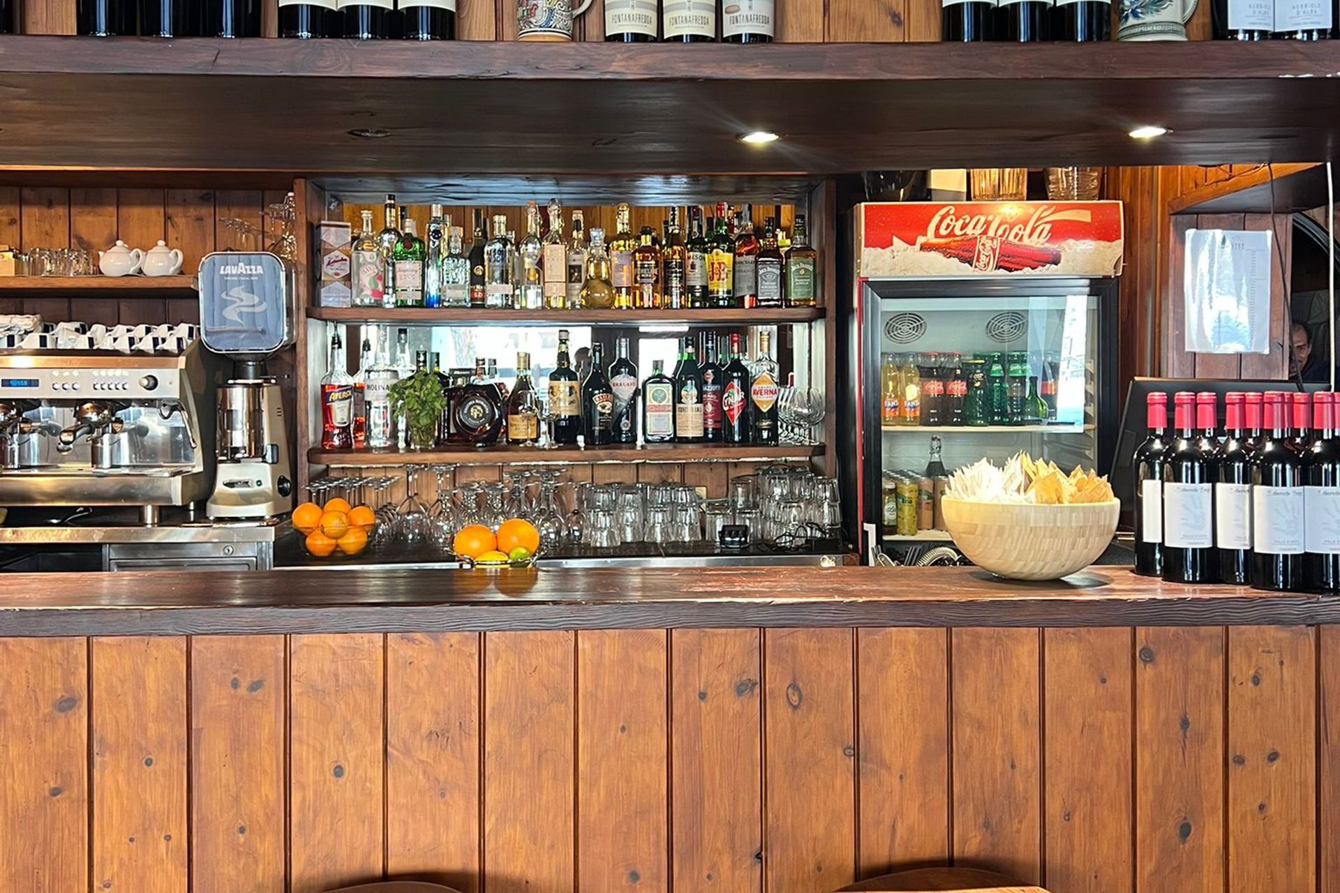 Bancone in legno con bottiglie di liquore, macchina per l'espresso, frigorifero per la Coca-Cola, bicchieri e arance.