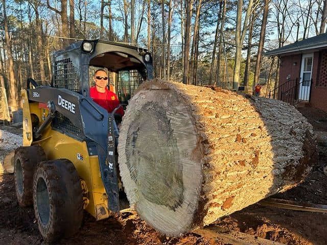A person in a yellow John Deere skid steer lifts a massive, thick-cut tree trunk in a residential yard.
