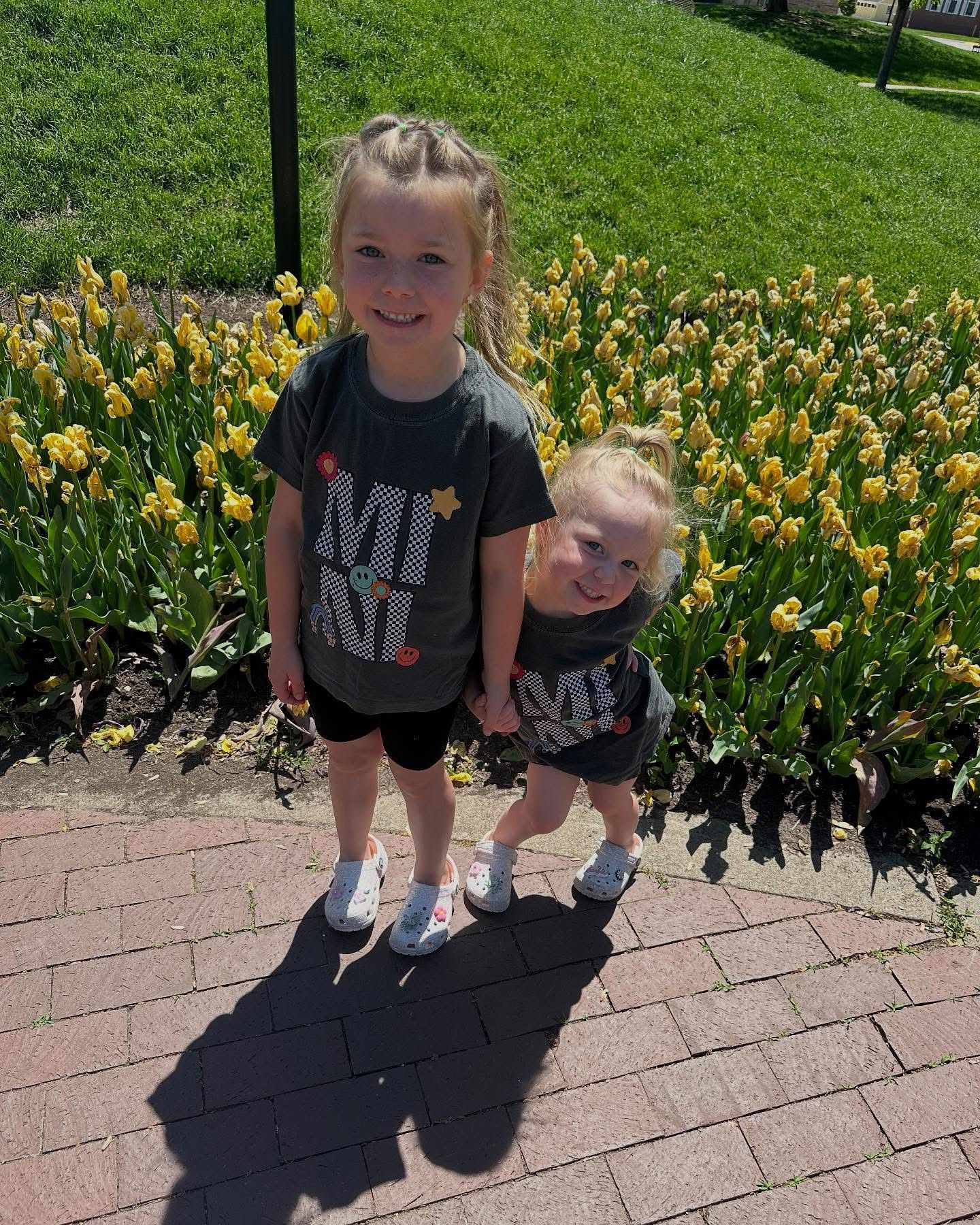 Two little girls are standing next to each other in front of a flower bed.
