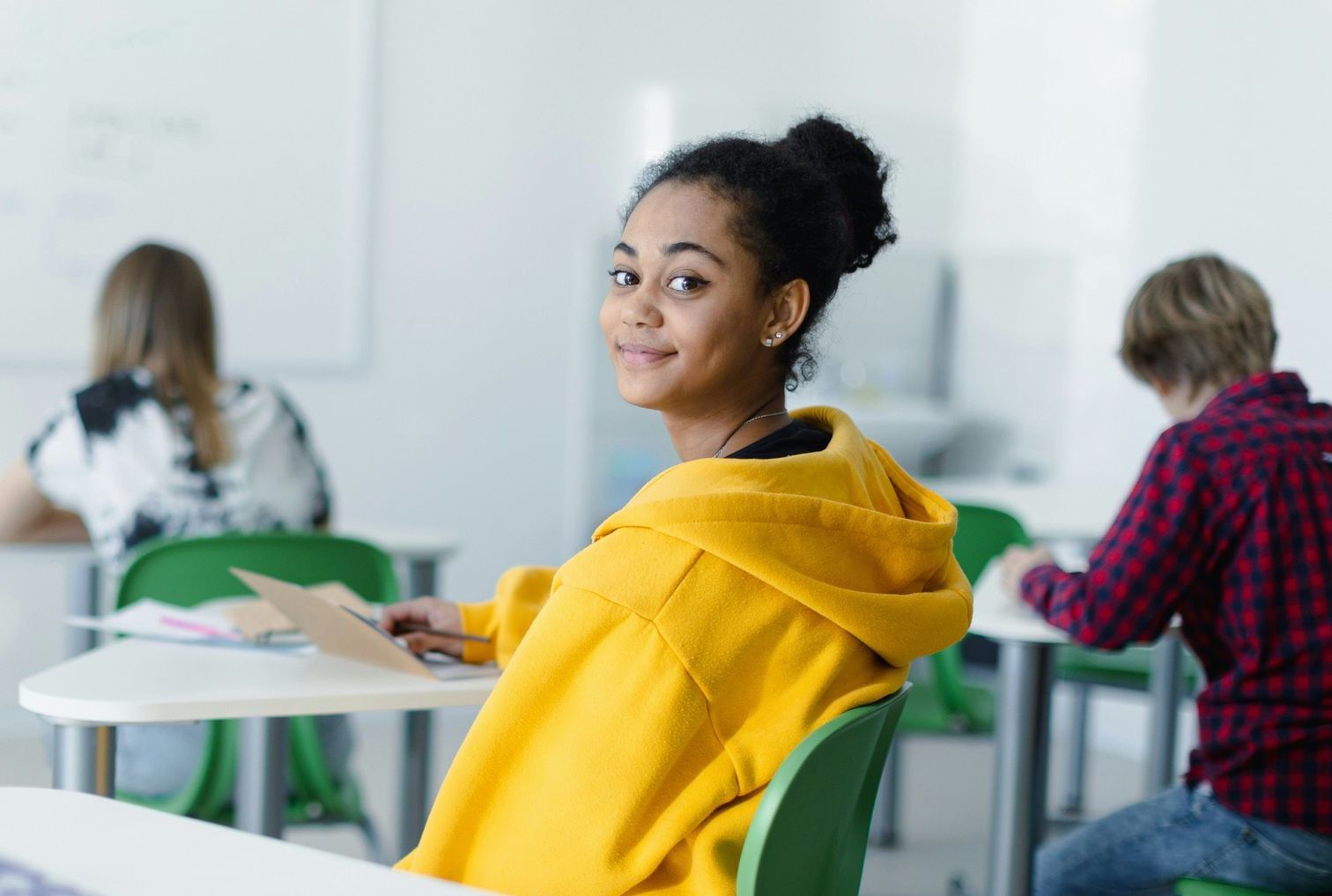 A girl in a yellow hoodie is sitting at a desk in a classroom.