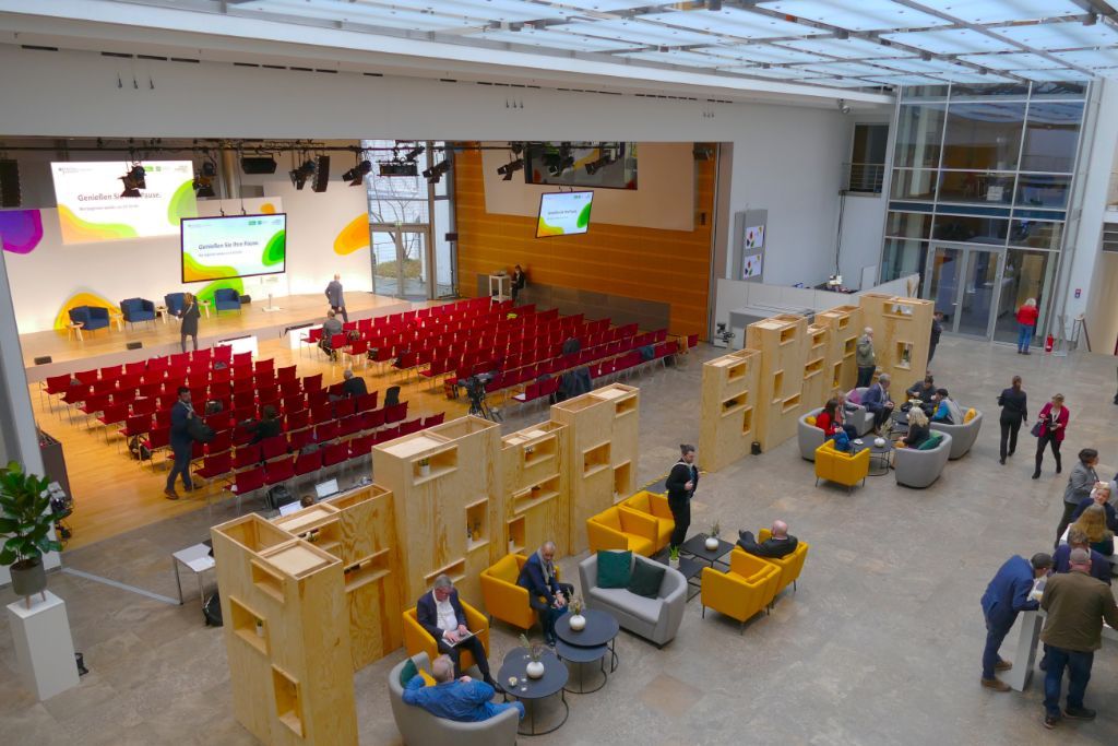 Großer Saal zweigeteilt. Rote Konferenzbestuhlung, Bühne, hölzerne Raumteiler, Lounge mit grauen und gelben Polstermöbeln