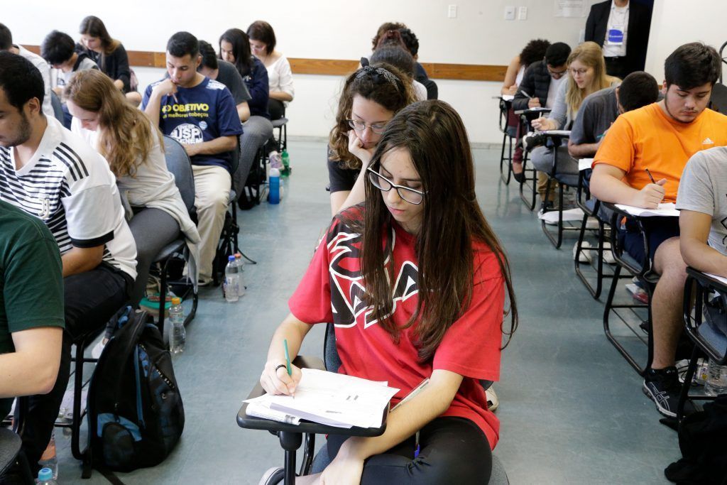 Sala cheia de estudantes realizando prova em carteiras escolares, concentrados e escrevendo.