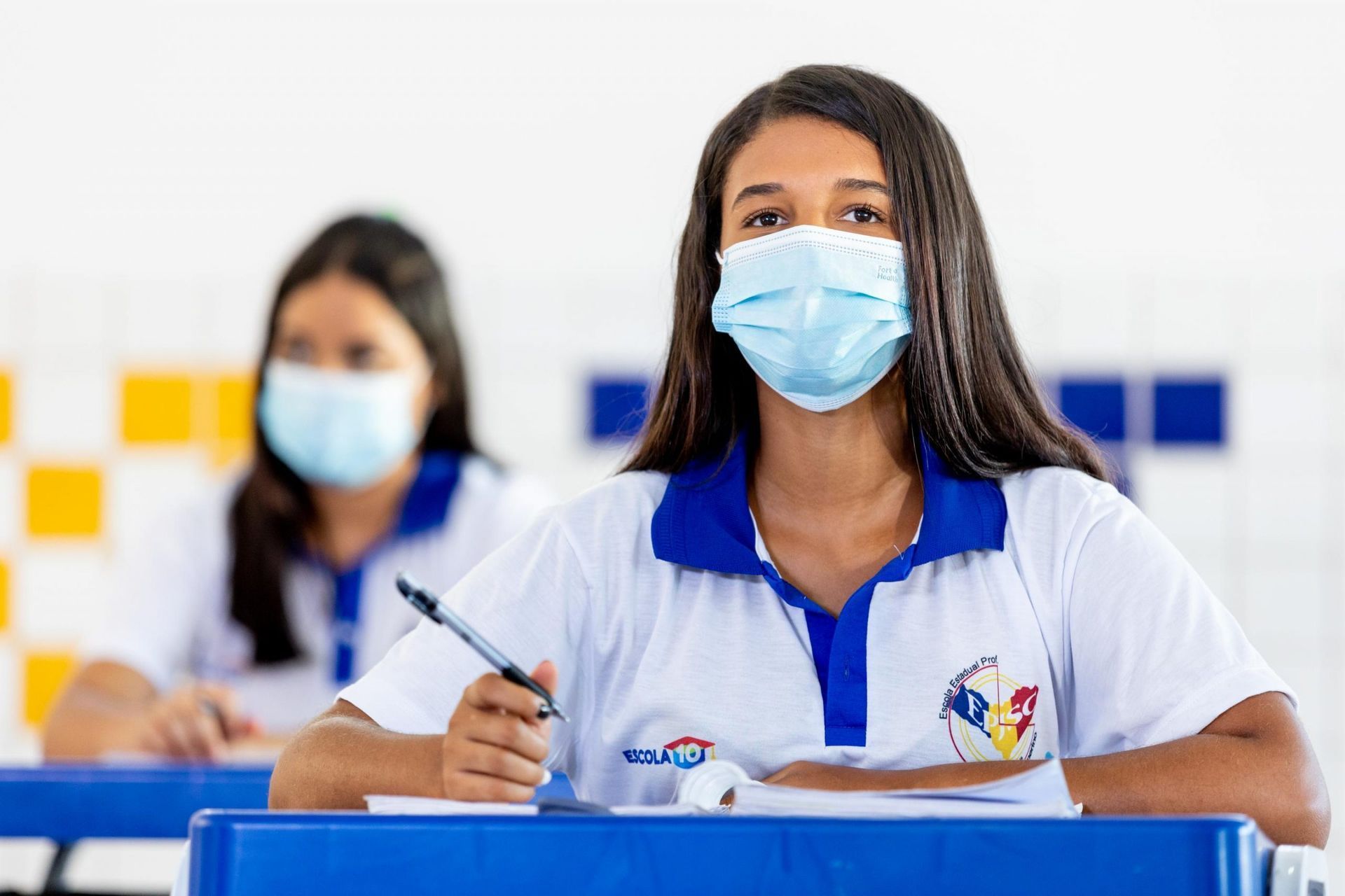Estudante usa máscara e participa ativamente da aula em sala com carteiras azuis e uniforme escolar.