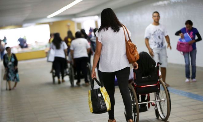 Mulher empurra jovem em cadeira de rodas por corredor público com outras pessoas ao fundo.
