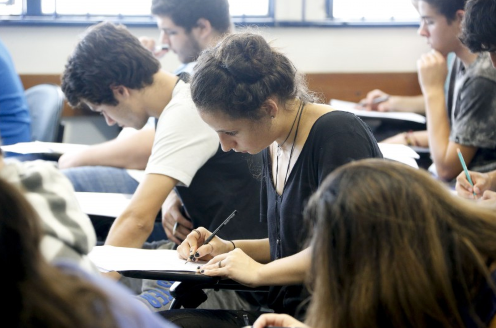 Estudantes em sala de aula realizando prova, concentrados e escrevendo em suas carteiras.