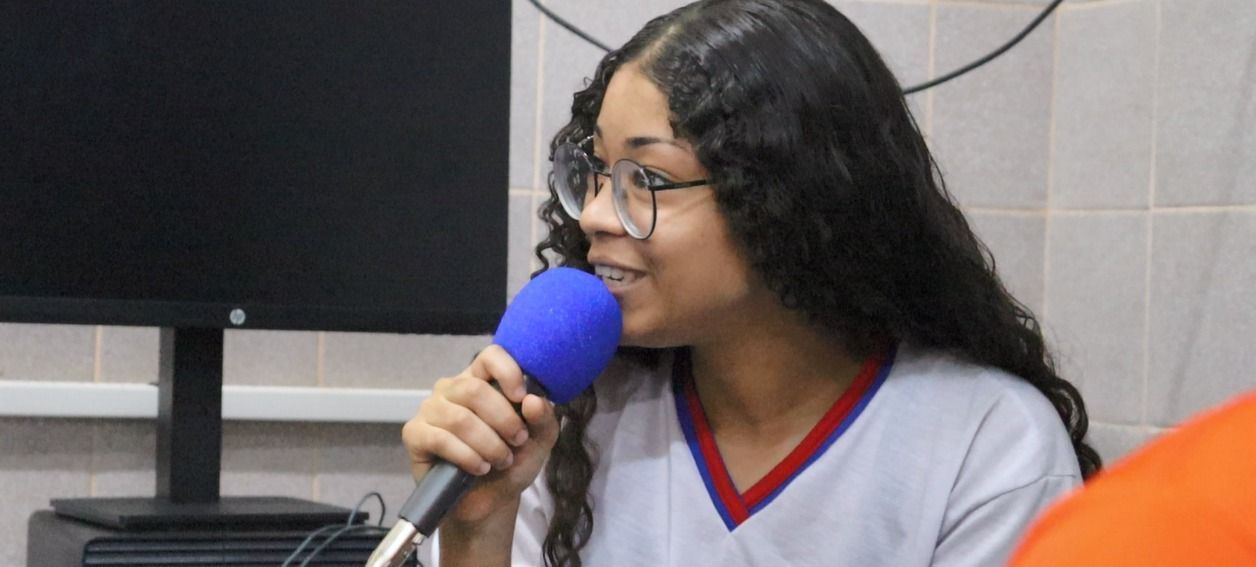 Jovem estudante com óculos fala ao microfone azul durante gravação em estúdio escolar