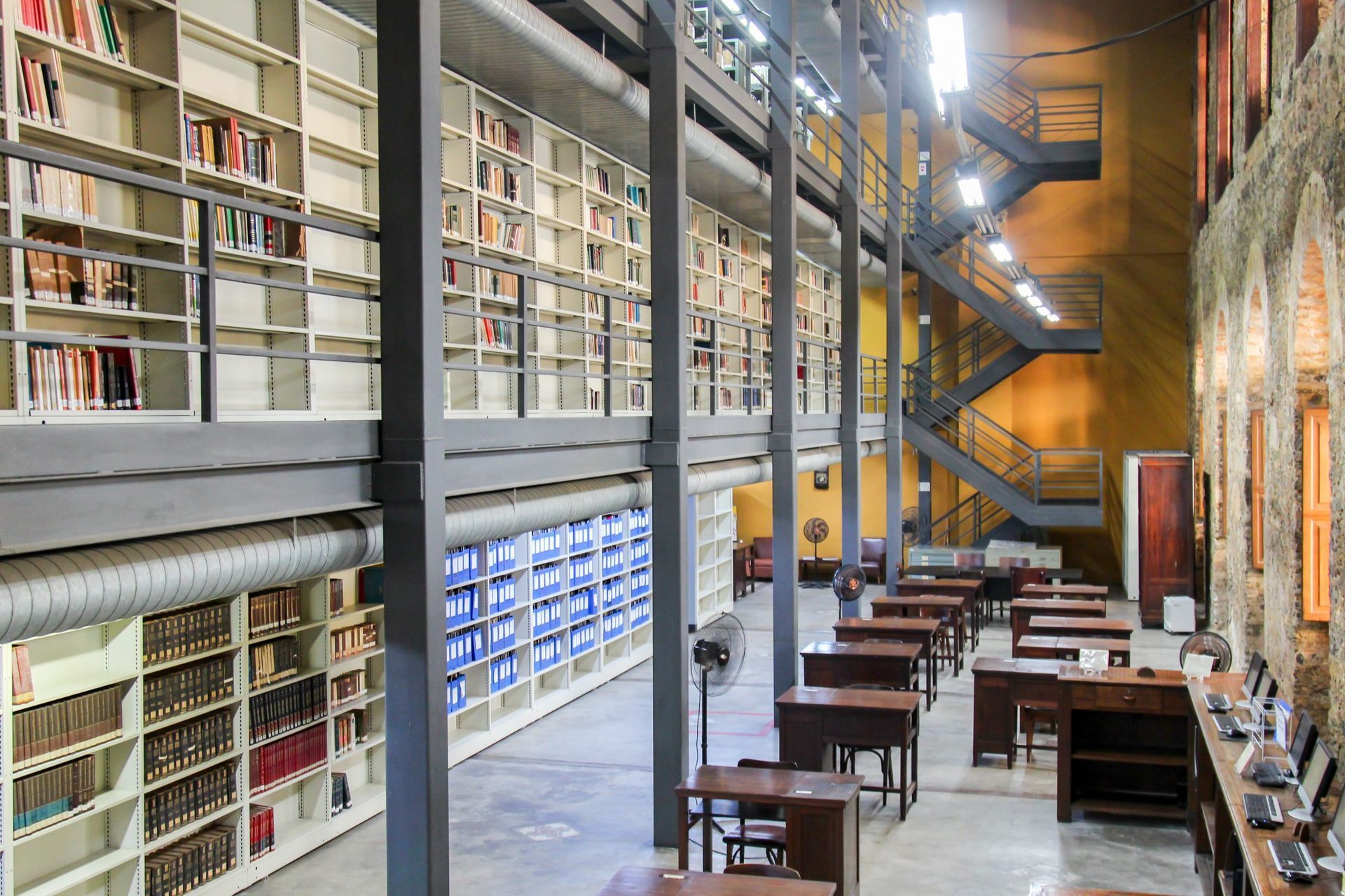 Biblioteca ampla com estantes altas, mesas de estudo e escadarias metálicas em estrutura industrial.