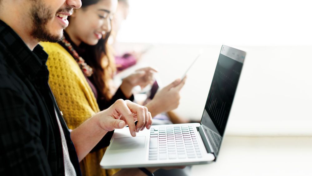 Jovens sentados usando notebook e tablet em ambiente claro, sorrindo e concentrados.