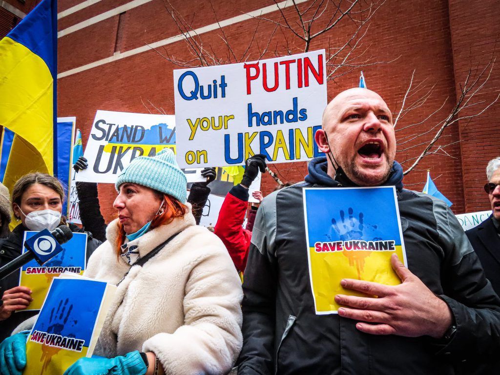 Manifestantes seguram cartazes “Quit PUTIN your hands on UKRAINE” e gritam em protesto pró-Ucrânia.