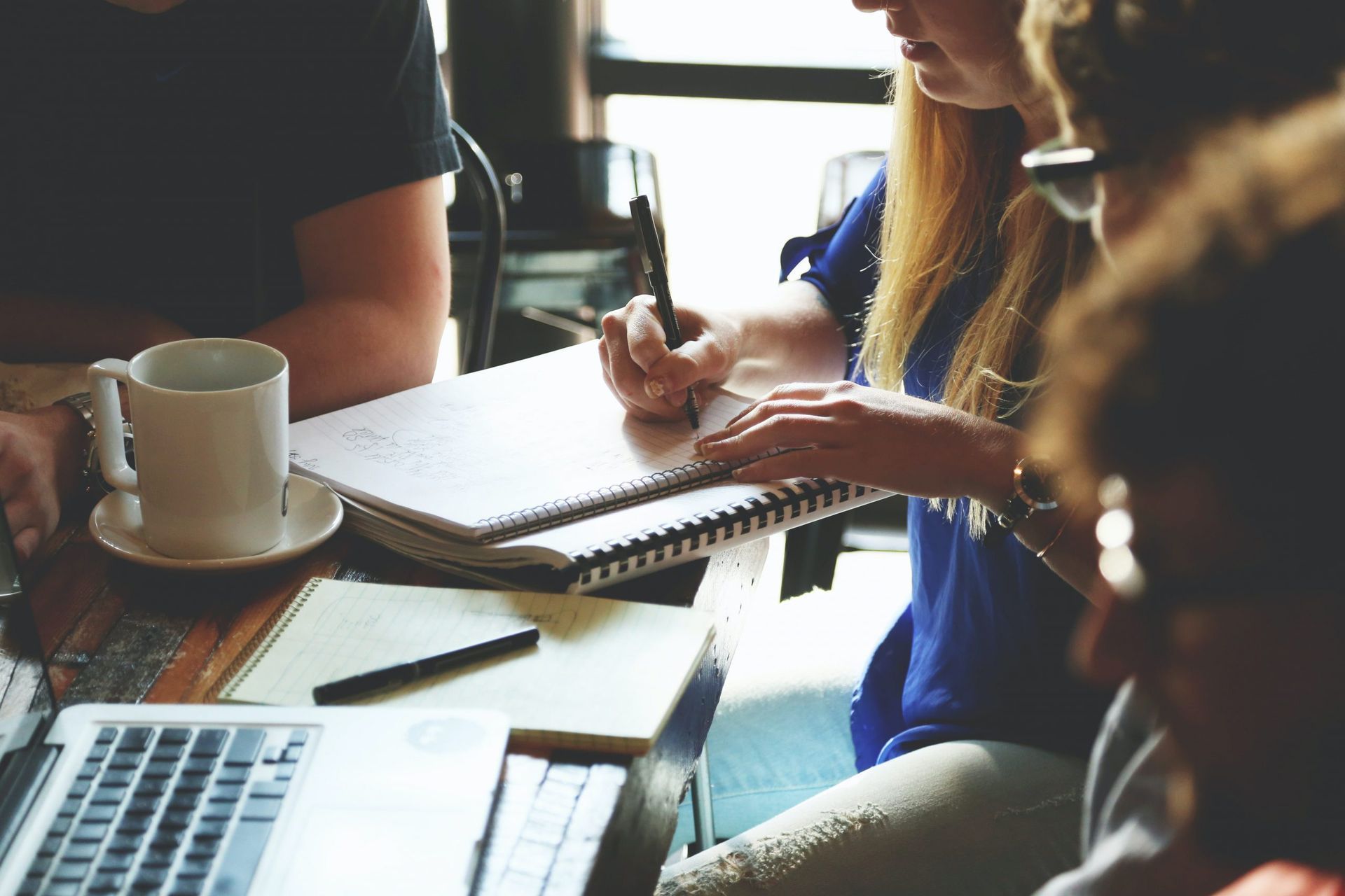 Grupo de estudantes fazendo anotações em cadernos ao redor de uma mesa com notebook e café.