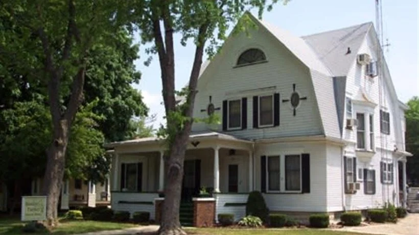 A large white house with black shutters is surrounded by trees.