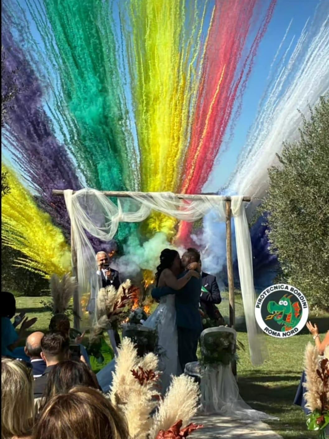 Coppia si bacia sotto festoni color arcobaleno durante una cerimonia nuziale all'aperto. Gli ospiti applaudono.