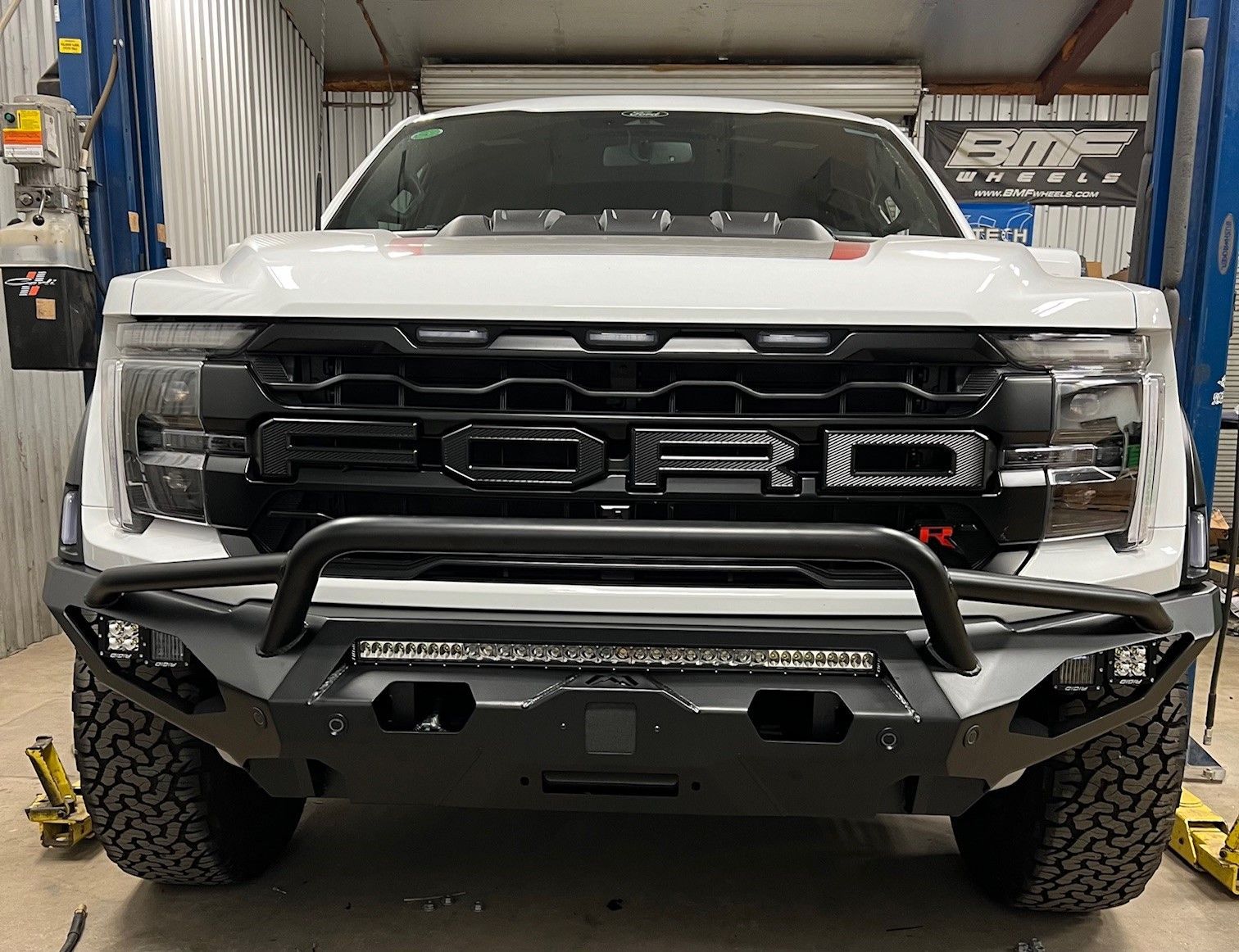 A white ford raptor with a black bumper is parked in a garage.