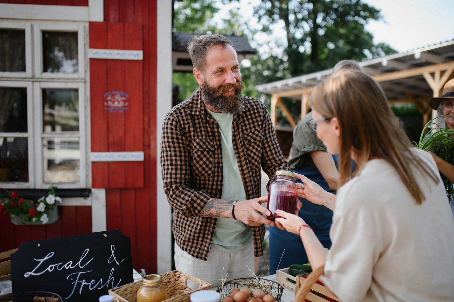 A bearded man hands a jar of preserves to a woman at a farmer's market, red building in the background.