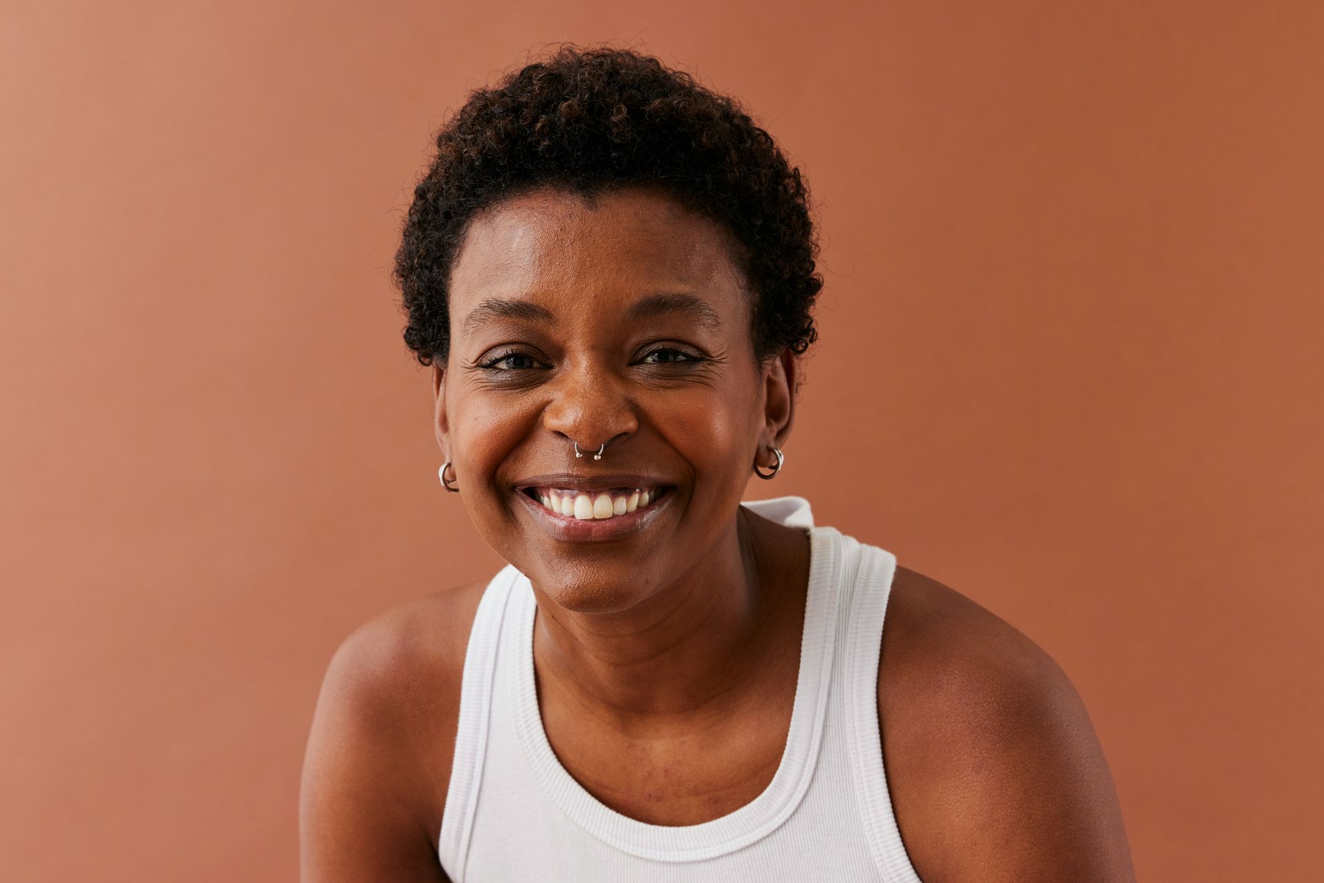 Woman with short, dark hair, wearing a white tank top, smiling, against a brown background.