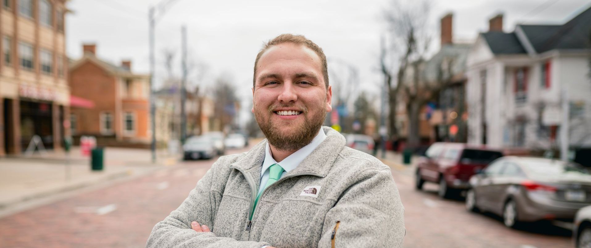 A man with a beard smiles in a street, arms crossed. He wears a sweater and tie. Buildings and cars in the background.