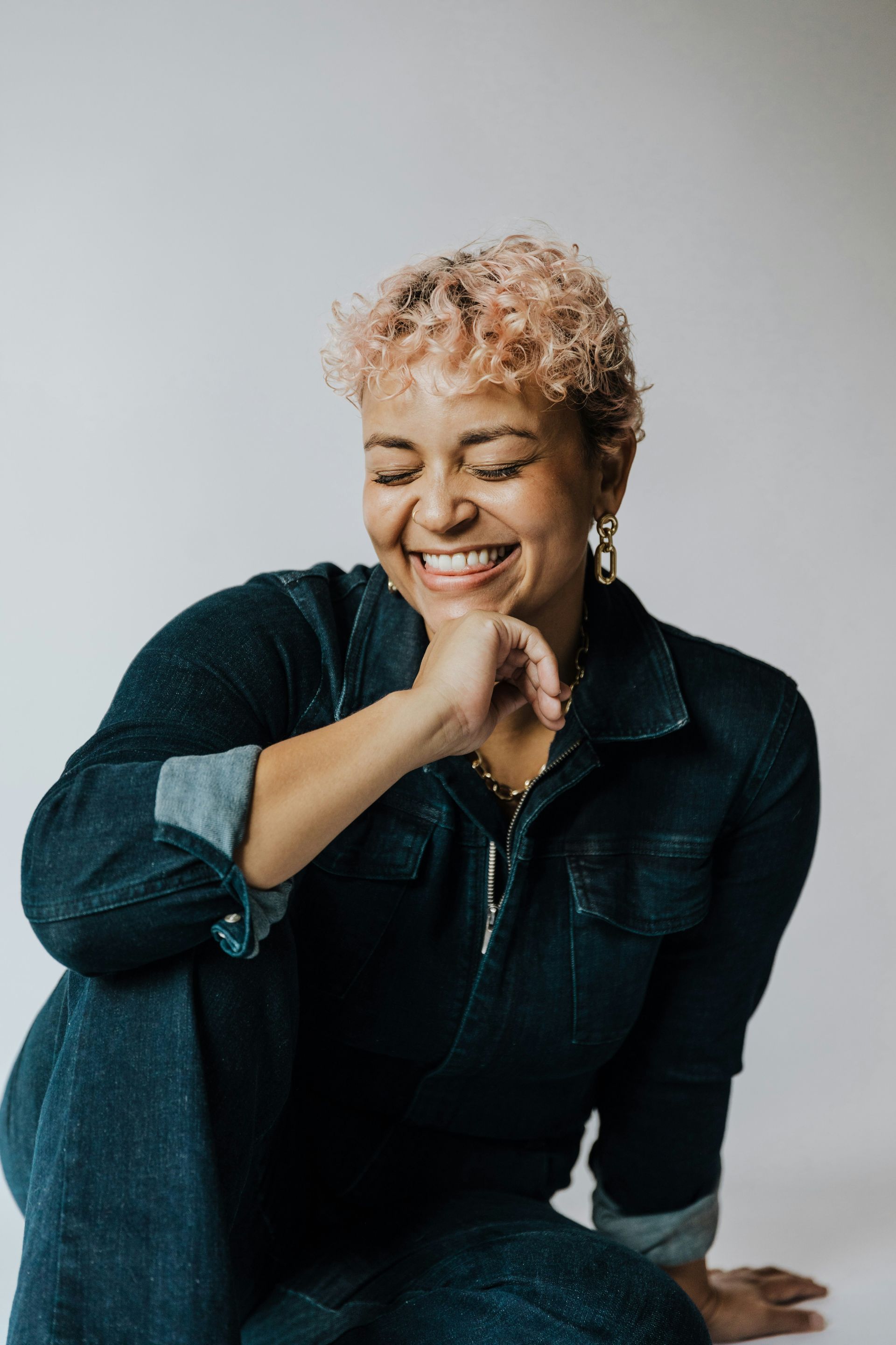Woman with pink curly hair, laughing, wearing a denim jumpsuit, sitting.