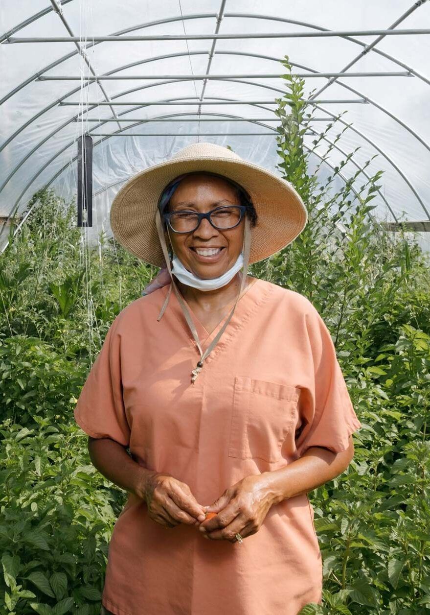 Woman in hat and scrubs smiles in a greenhouse, holding a tomato; green plants surround her.