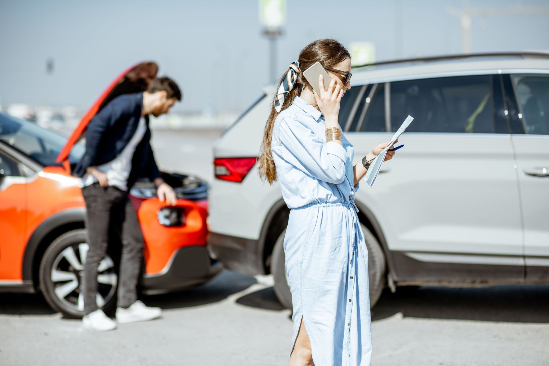 A woman in a blue dress talks on her phone while a man checks the engine of his orange car after a traffic accident. A woman in a blue dress talks on her phone while a man checks the engine of his orange car after a traffic accident.
