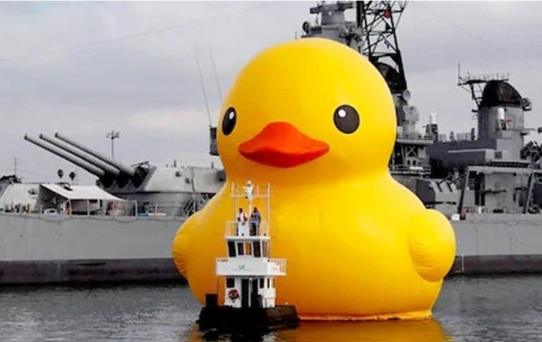 Giant yellow inflatable rubber duck floats in water next to a tugboat, with a battleship in the background. Giant yellow inflatable rubber duck floats in water next to a tugboat, with a battleship in the background.