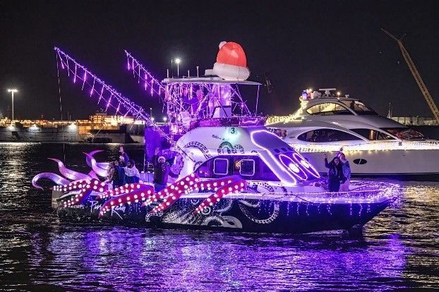 Festive boat parade with a boat decorated as an octopus in purple lights, wearing a Santa hat.