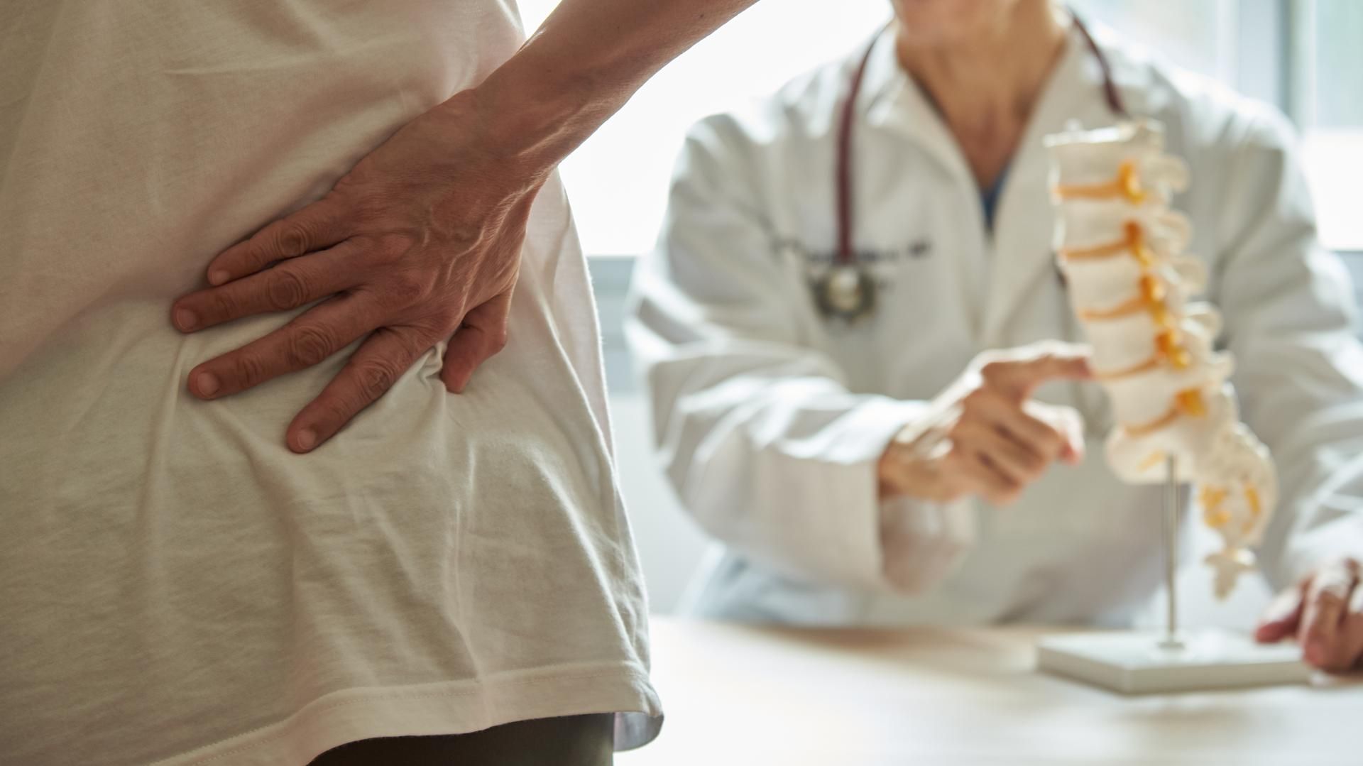 A woman is holding her back while a doctor shows her a model of a spine.