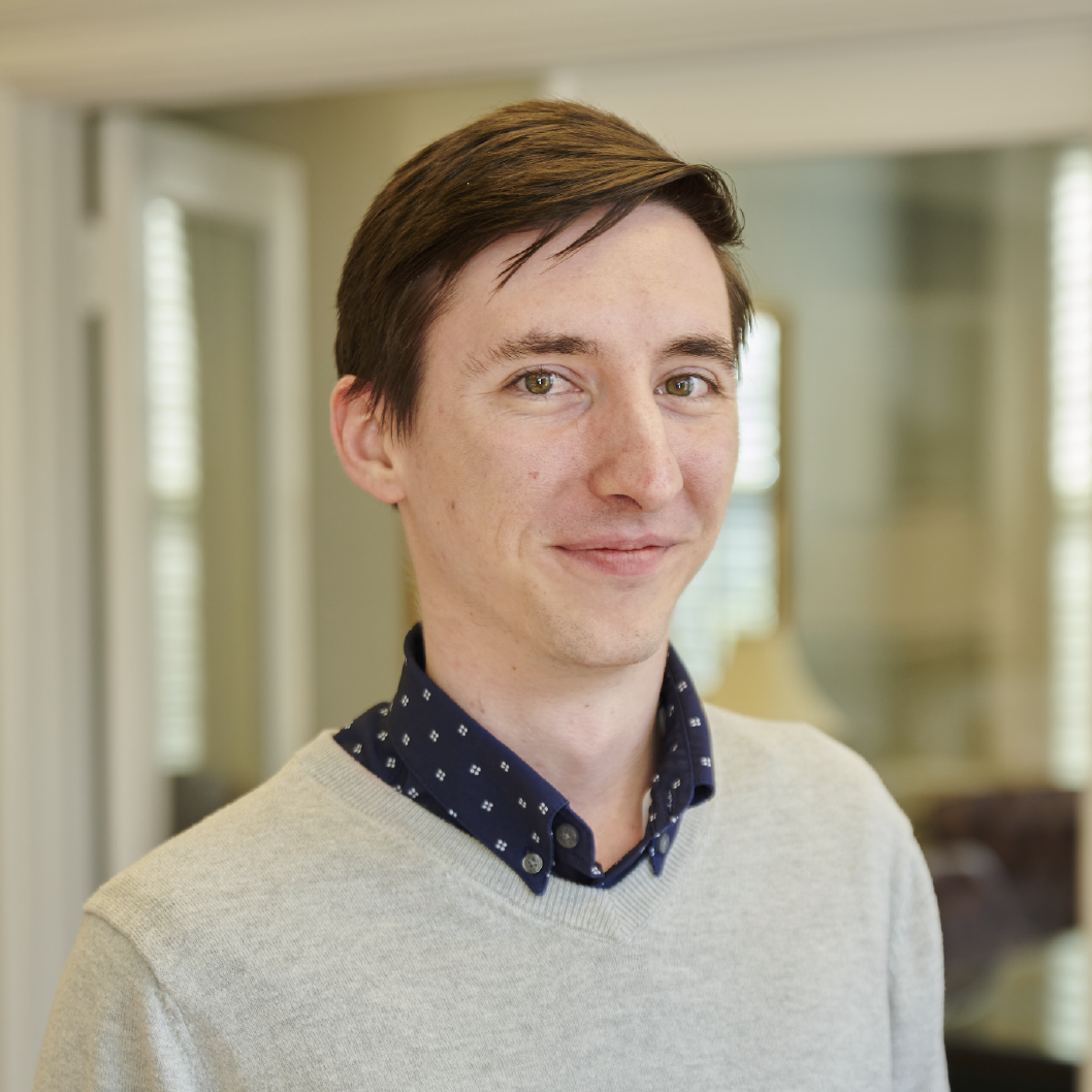 A man wearing a grey sweater and a blue polka dot shirt smiles for the camera