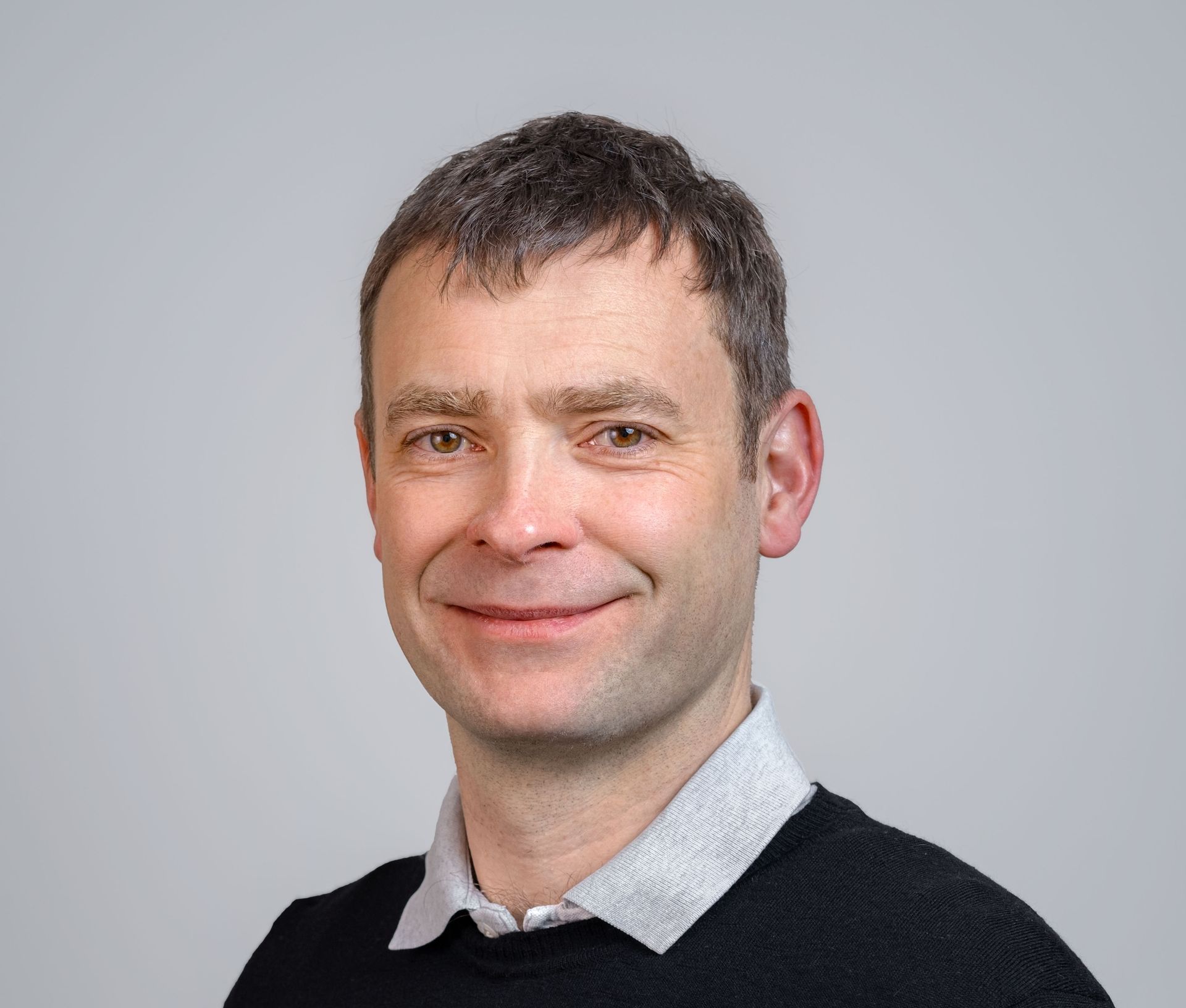 Man with short brown hair, smiling, wearing a black sweater over a collared shirt, against a grey background.