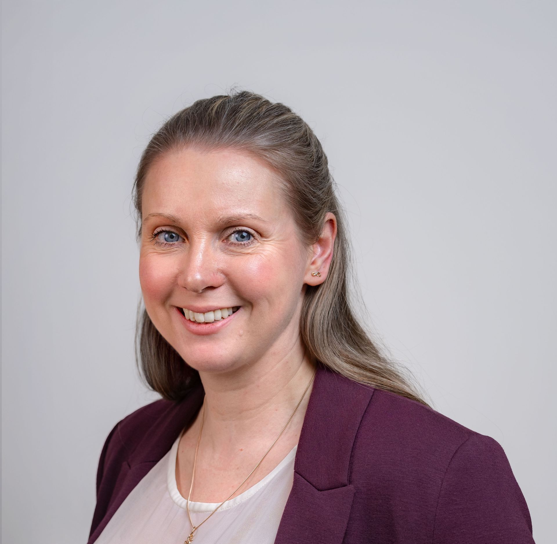 Woman with light skin, smiling, wearing a pink blouse and burgundy blazer, against a light background.