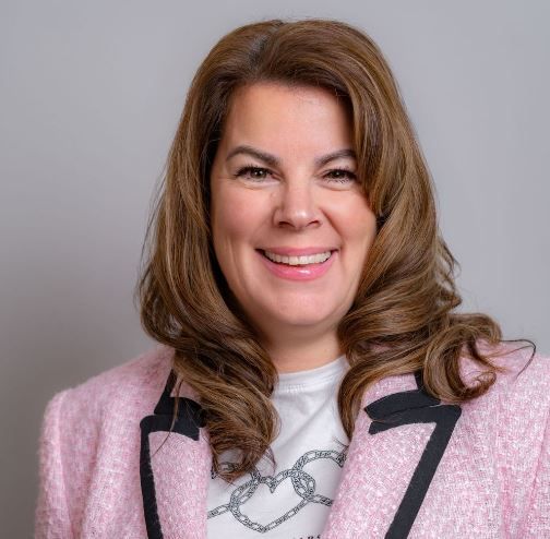 Woman with brown hair smiles in a pink jacket and white shirt. Gray background.