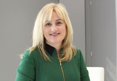 Blond woman in green jacket smiles. Background is gray wall and white square.