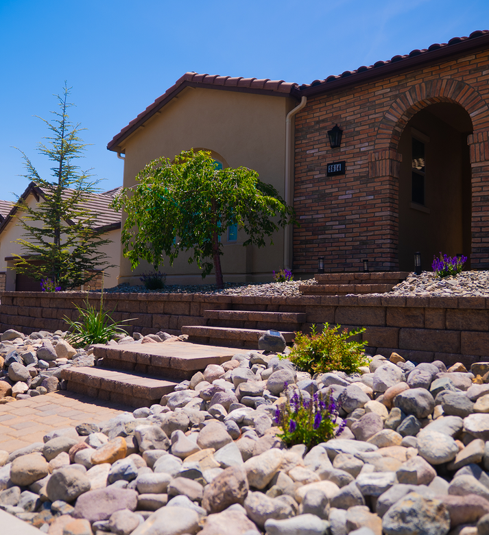 Tan house with brick facade, stone steps, and landscaping, under a blue sky.