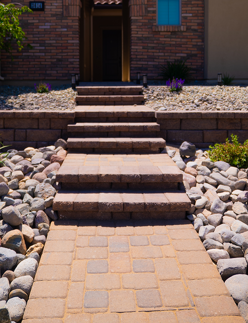 Brick pathway leading to a home's front door, with steps, landscaping, and a blue window.