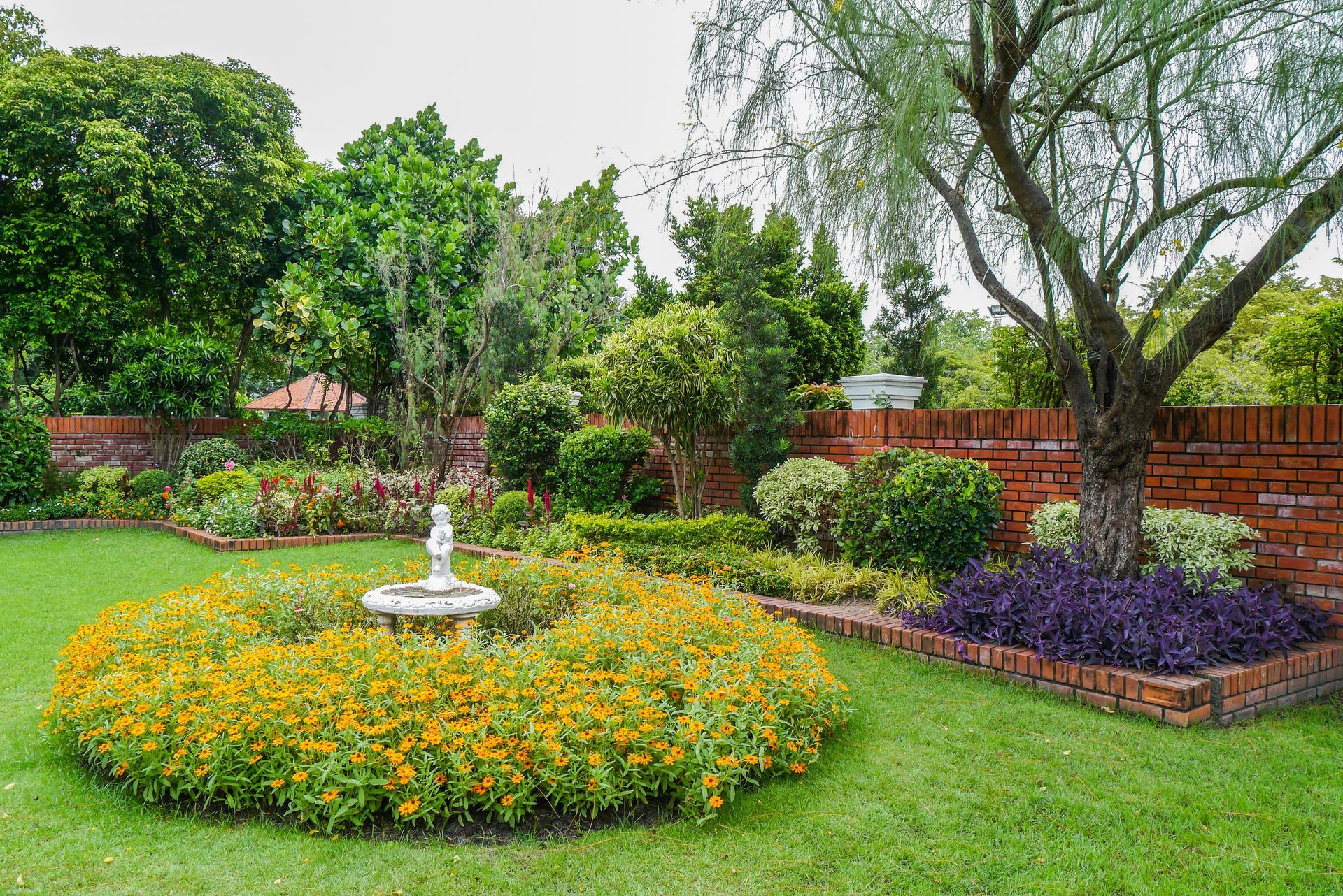 Lush garden with a circular flowerbed around a fountain, surrounded by green grass, trees, and a brick wall.