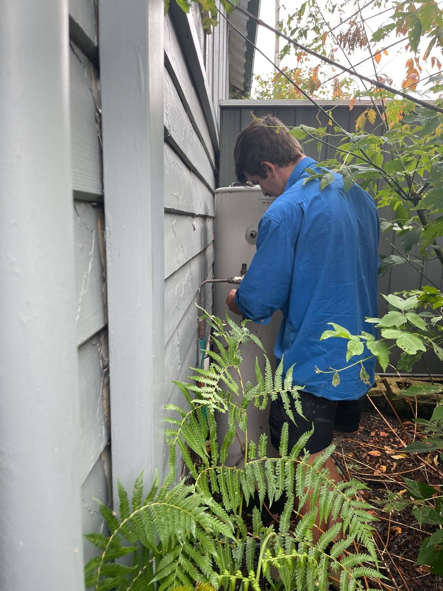Man in Blue Shirt Working on a Water Heater Outside a House — Expert Plumbing & Solar Services Bathurst in Kelso, NSW