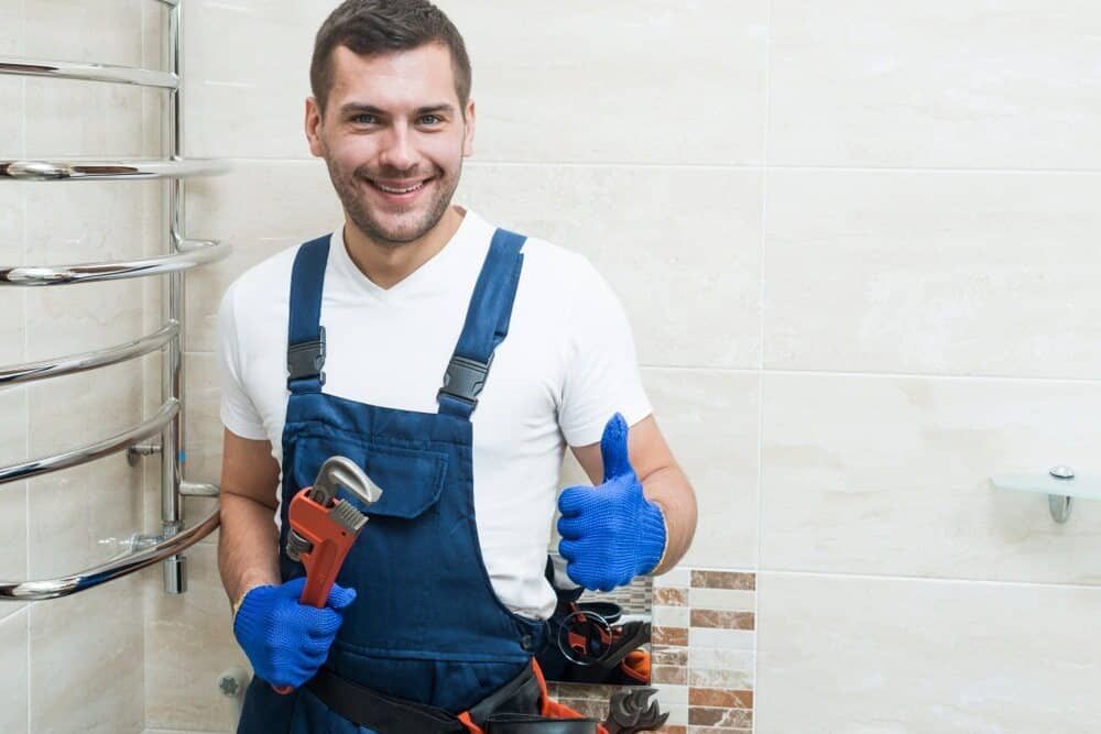 Plumber in Blue Overalls Gives a Thumbs Up While Holding a Wrench in a Bathroom — Expert Plumbing & Solar Services Bathurst in Blayney, NSW