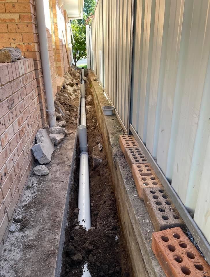 Trench With White Pipe Between a Brick Wall and Corrugated Metal Fence — Expert Plumbing & Solar Services Bathurst in Kelso, NSW