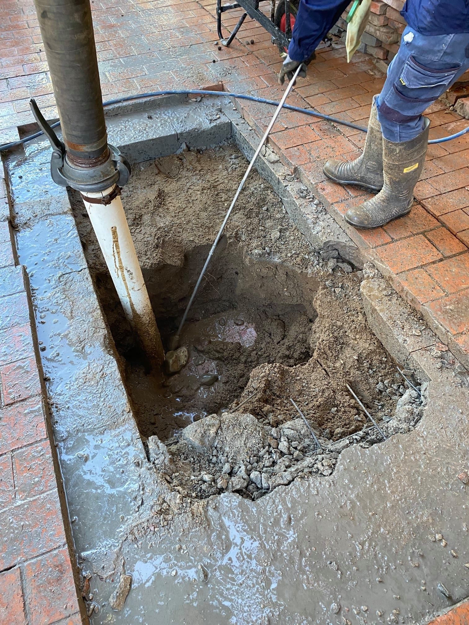 a Person in Boots Probes a Rectangular Hole Around a White Pipe Set in Concrete — Expert Plumbing & Solar Services Bathurst in Kelso, NSW