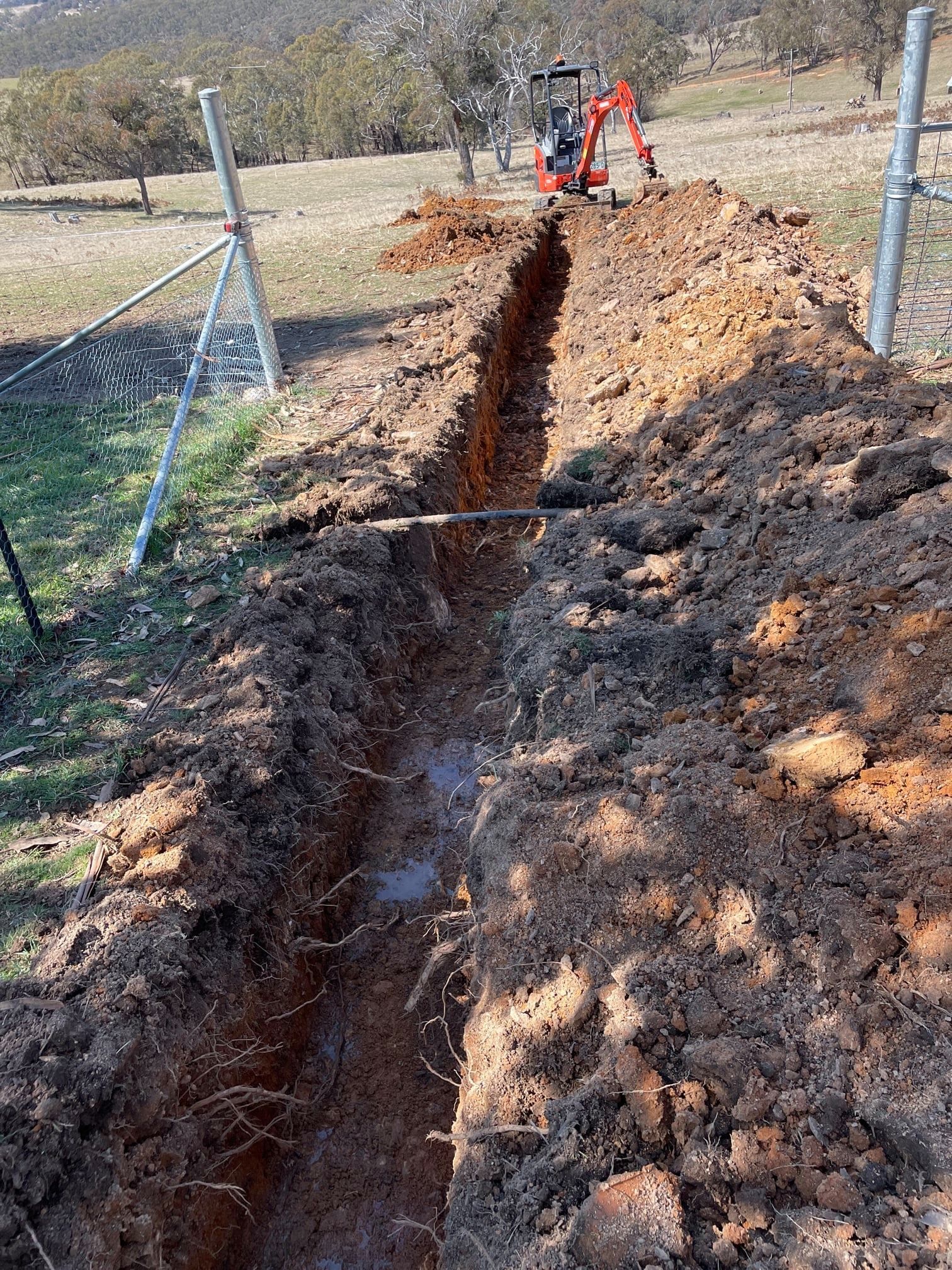 A Trench Being Dug by an Excavator Along a Fence Line in a Rural Setting — Expert Plumbing & Solar Services Bathurst in Orange NSW