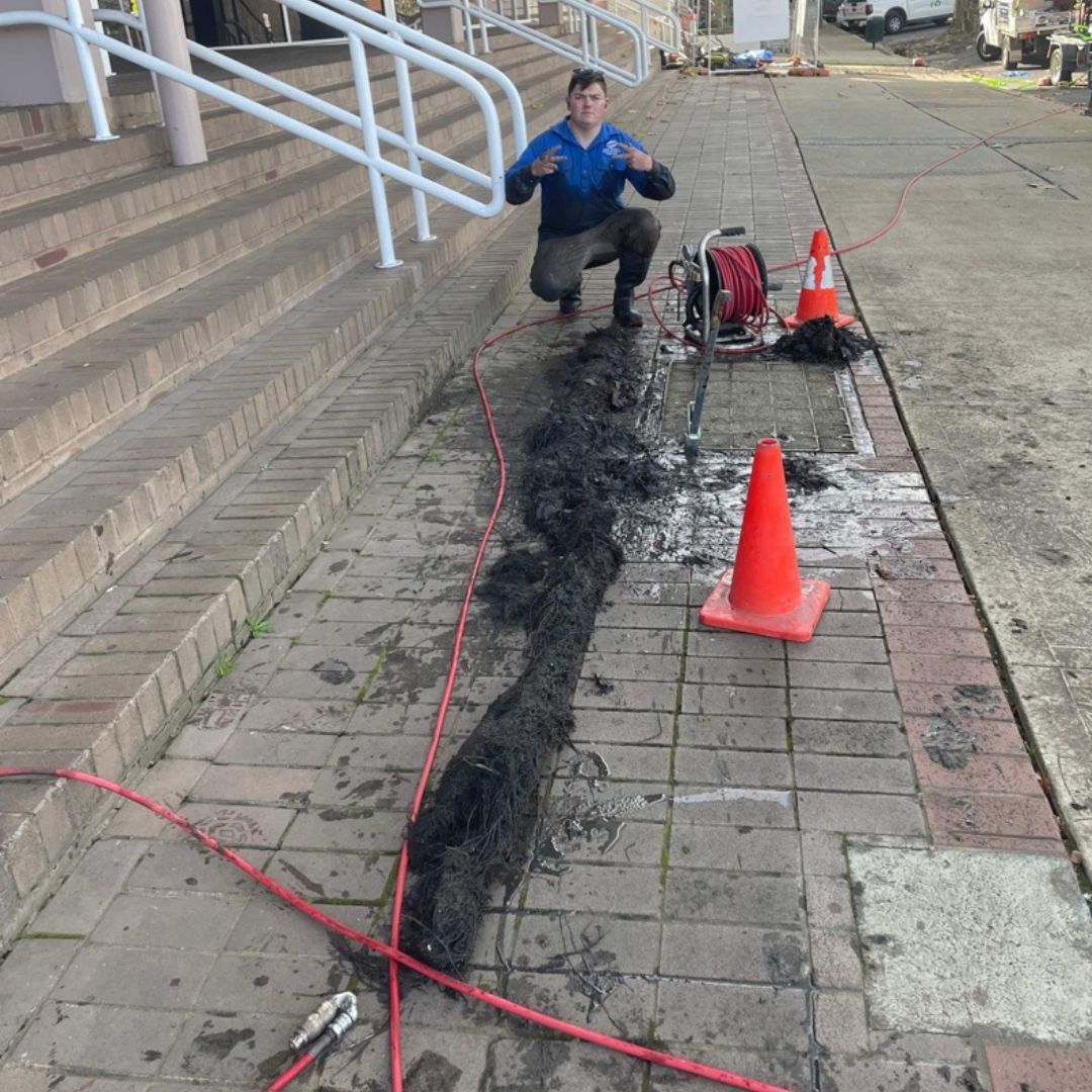 Man Kneels Next to a Long, Dark Hair Clog Extracted From a Drain — Expert Plumbing & Solar Services Bathurst in Kelso, NSW