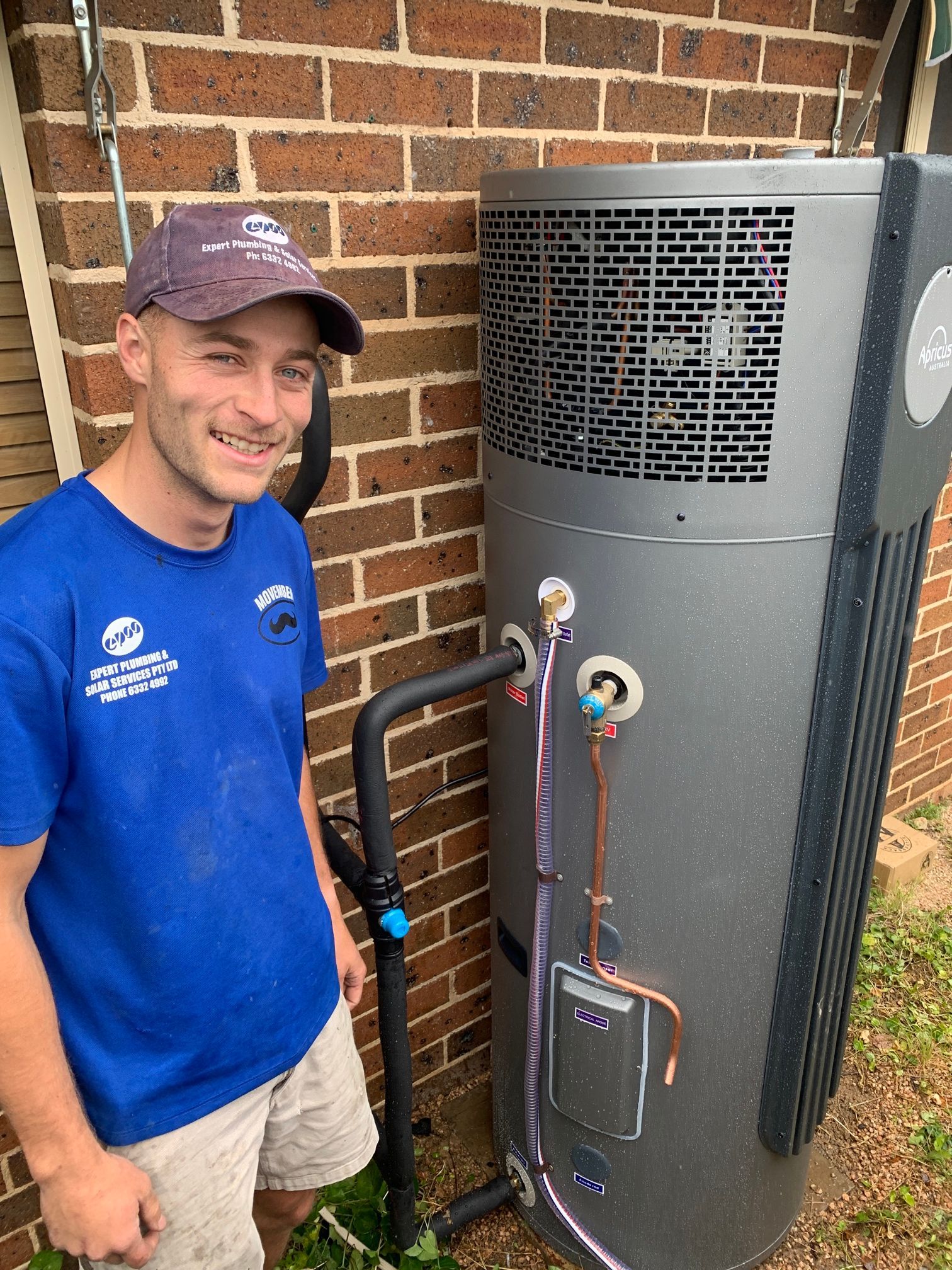 Man in Blue Shirt and Cap Stands Next to a Large Gray Water Heater — Expert Plumbing & Solar Services Bathurst in Kelso, NSW