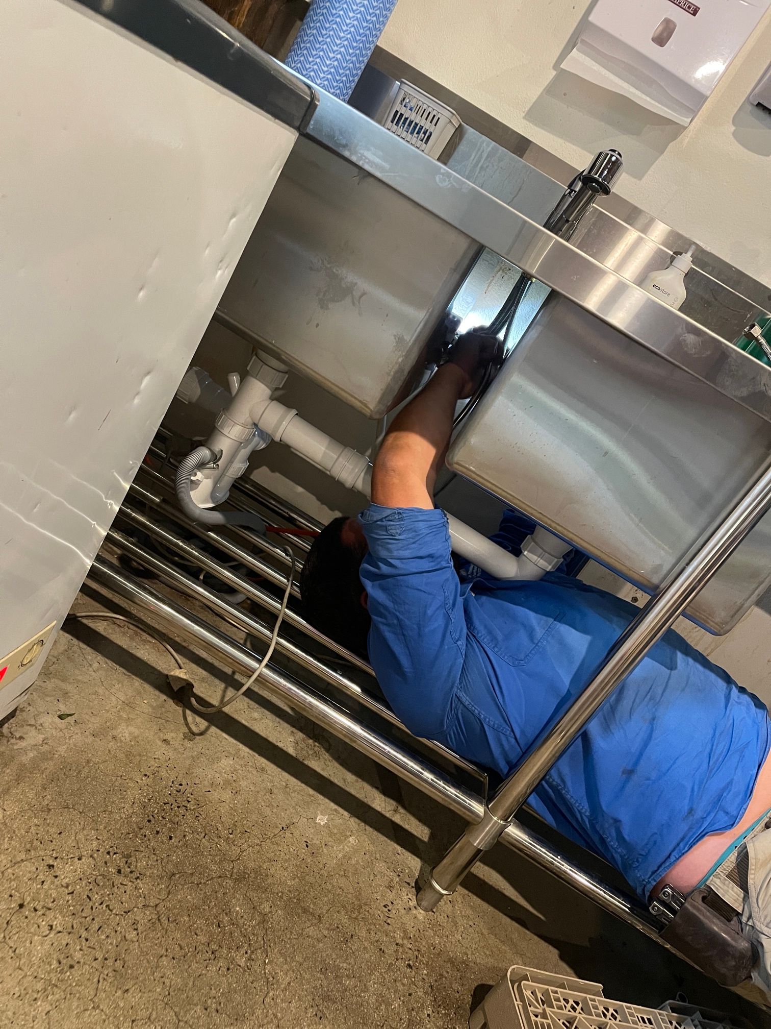 Plumber in Blue Shirt Works Under Stainless Steel Sink — Expert Plumbing & Solar Services Bathurst in Kelso, NSW