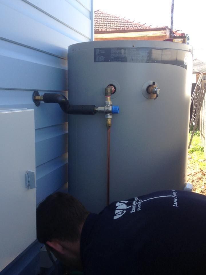 A Plumber Works on a Grey Hot Water Tank Next to a Blue Wall — Expert Plumbing & Solar Services Bathurst in Kelso, NSW