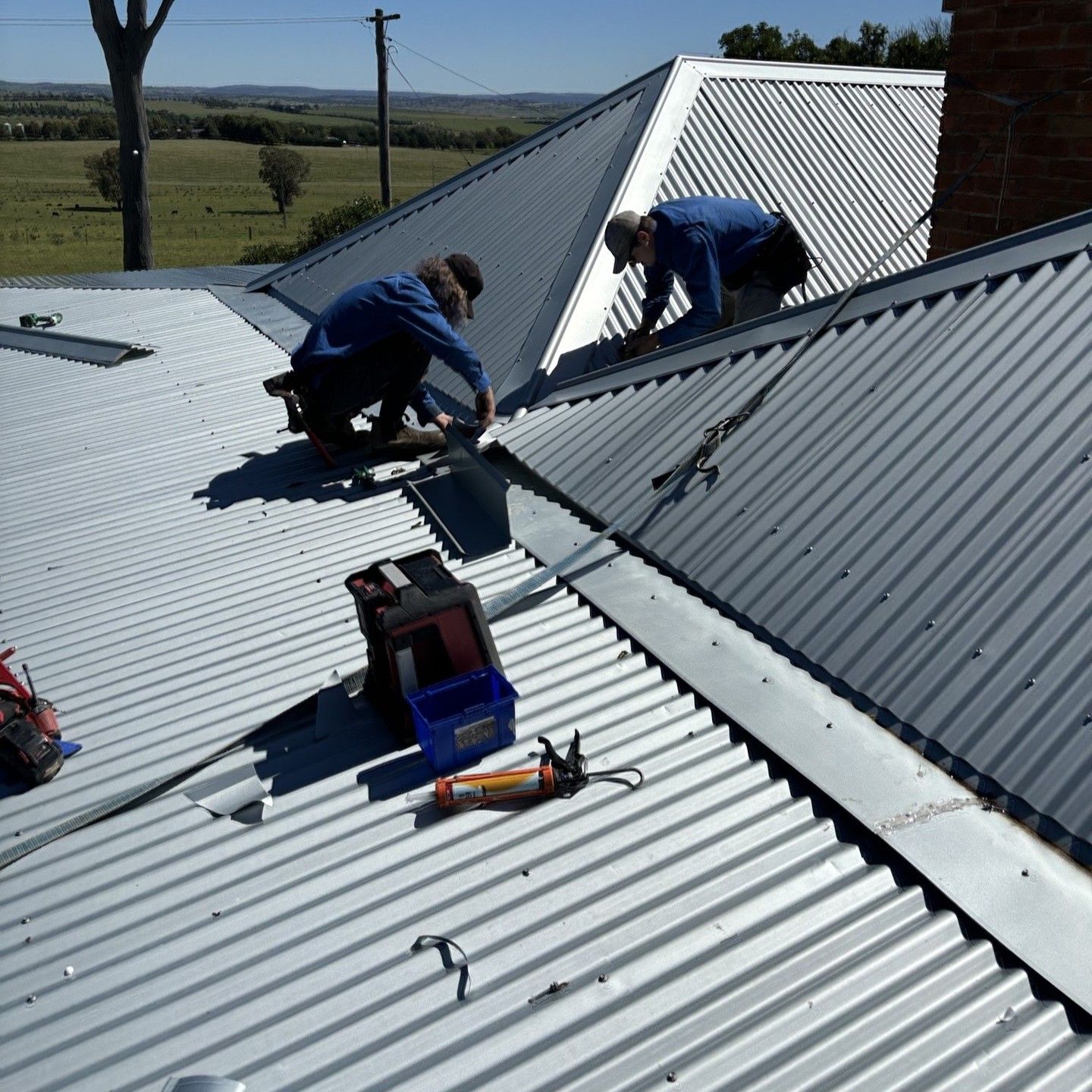 Two Workers in Blue Coveralls on a Corrugated Metal Roof — Expert Plumbing & Solar Services Bathurst in Lithgow, NSW