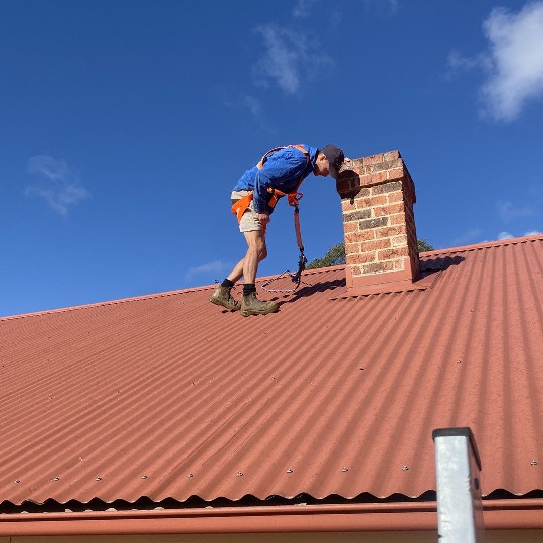 Person on a Red Corrugated Metal Roof, Examining a Brick Chimney — Expert Plumbing & Solar Services Bathurst in Lithgow, NSW