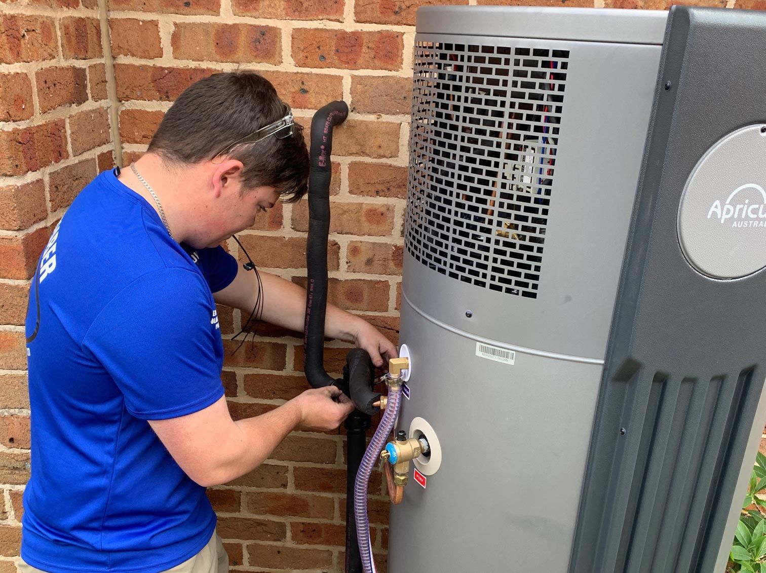 Technician in Blue Shirt Working on an Apricus Heat Pump Next to a Brick Wall — Expert Plumbing & Solar Services Bathurst in Kelso, NSW