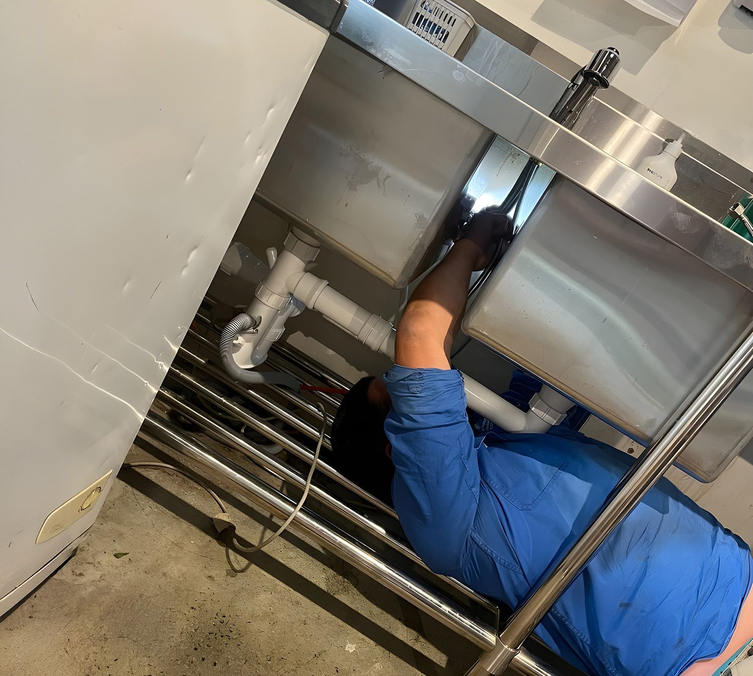 A Plumber In A Blue Shirt Lies On The Floor While Repairing Plumbing Pipes Under A Stainless-Steel K